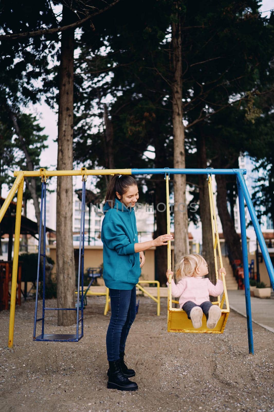 Mom swings a little girl on a swing in the park and looks at her. High quality photo