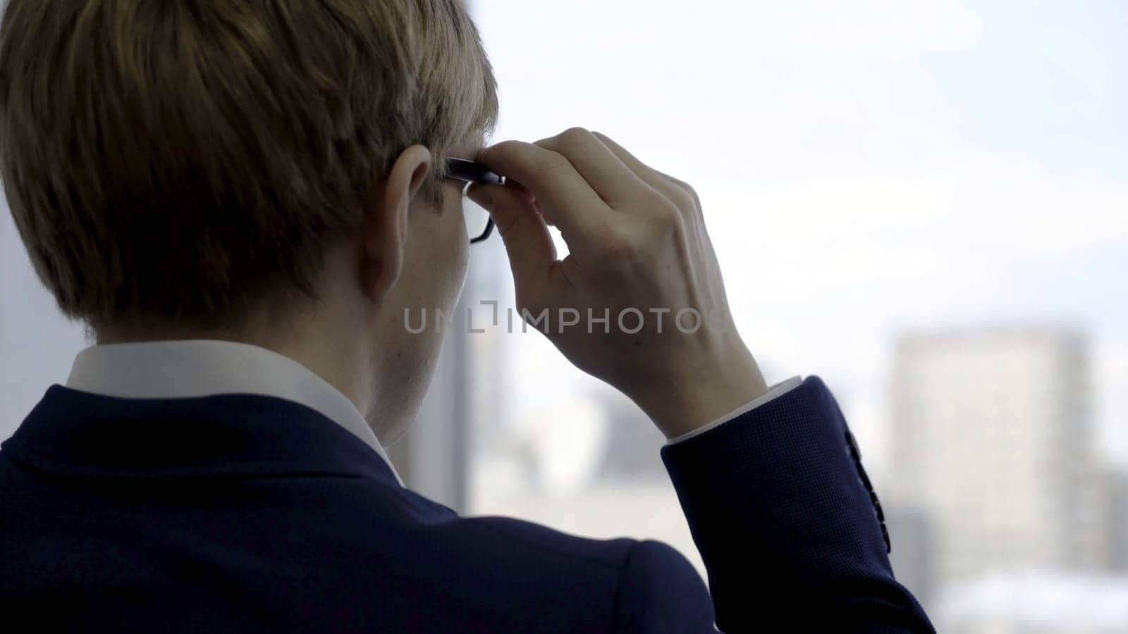 Back view of successful businessman in suit in the office looking out of the window and touching his glasses, close up. Rear view of a man looking through the window at the city