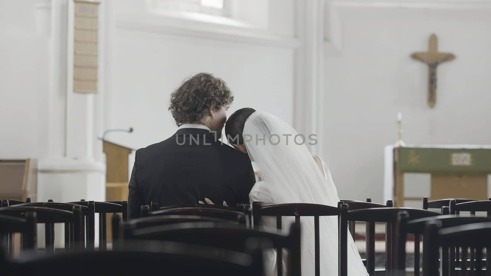 Newlyweds in love look at each other in church. Action. Beautiful newlyweds are sitting on chairs in church and looking at each other. Newlyweds in love watch and tenderly embrace while sitting in church.