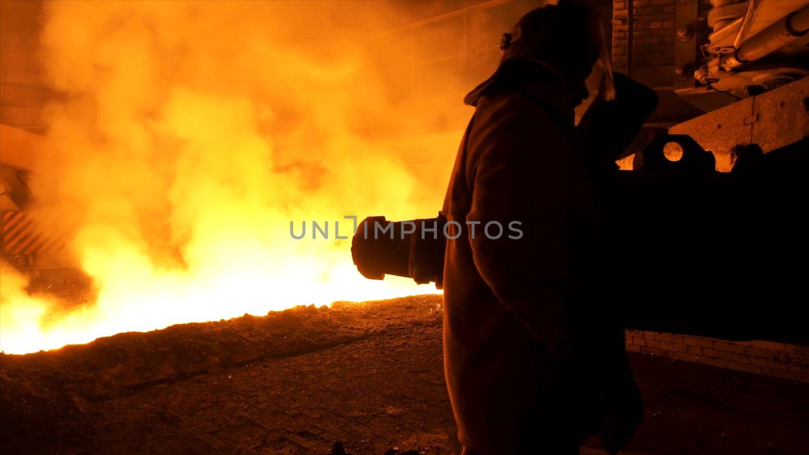 Man worker in special protective clothing standing near molten steel with sparkles and steam, heavy industry concept. Molten metal flowing in chute at the factory