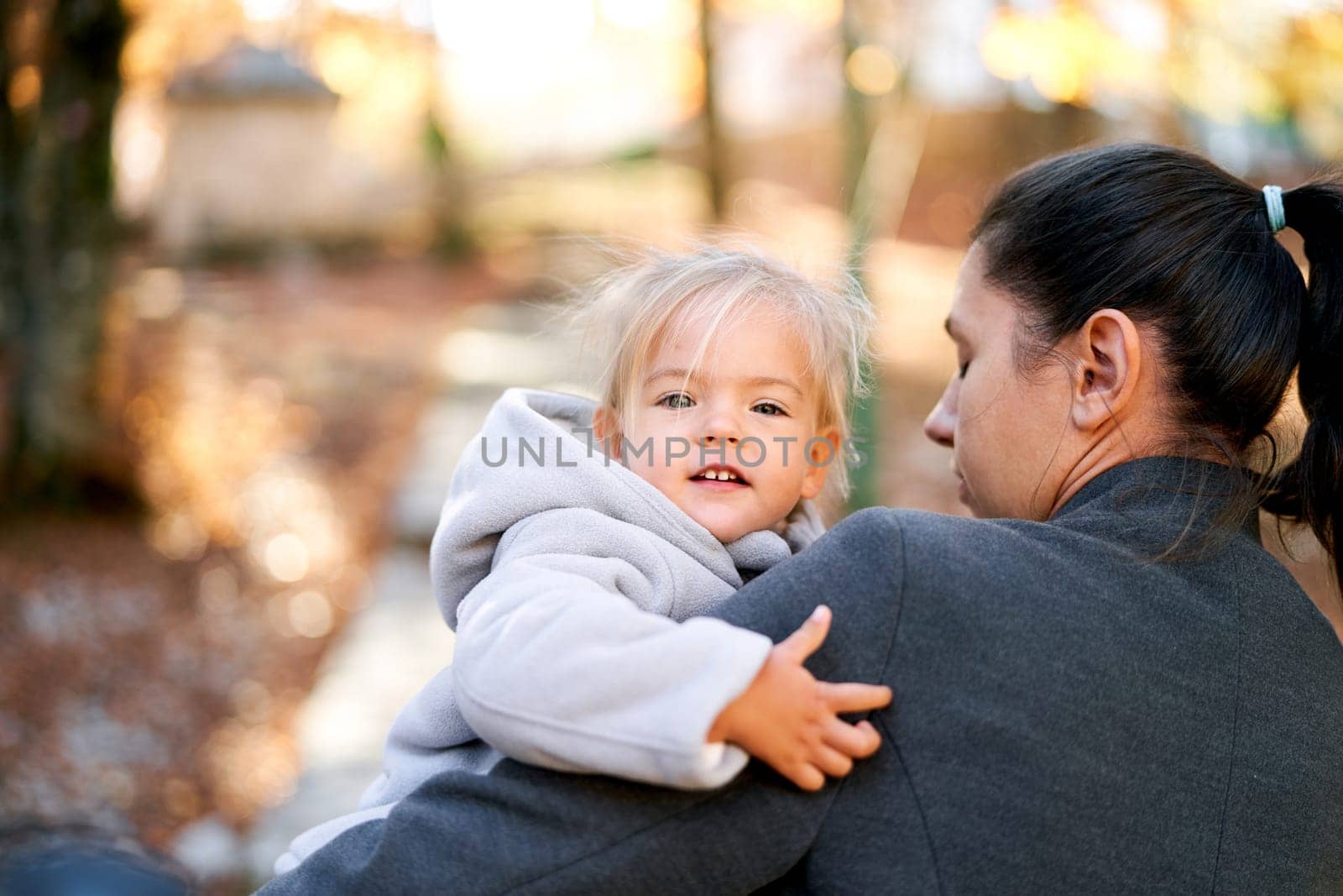 Smiling little girl peeking over her mother shoulder while sitting in her arms. High quality photo