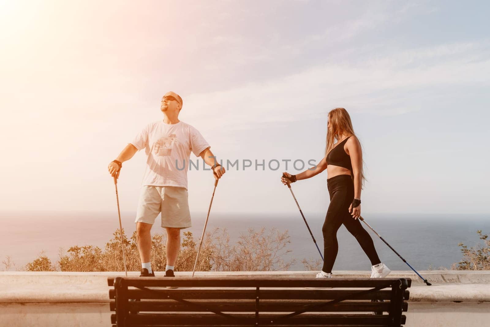 Happy Middle aged couple or friends practicing nordic walking in park near sea. Mature couple with trekking poles walking, practicing Nordic walking outdoors. Aging youthfully and sport concept by panophotograph