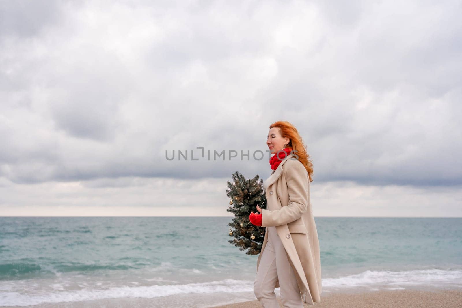 Redhead woman Christmas tree sea. Christmas portrait of a happy redhead woman walking along the beach and holding a Christmas tree in her hands. Dressed in a light coat, white suit and red mittens