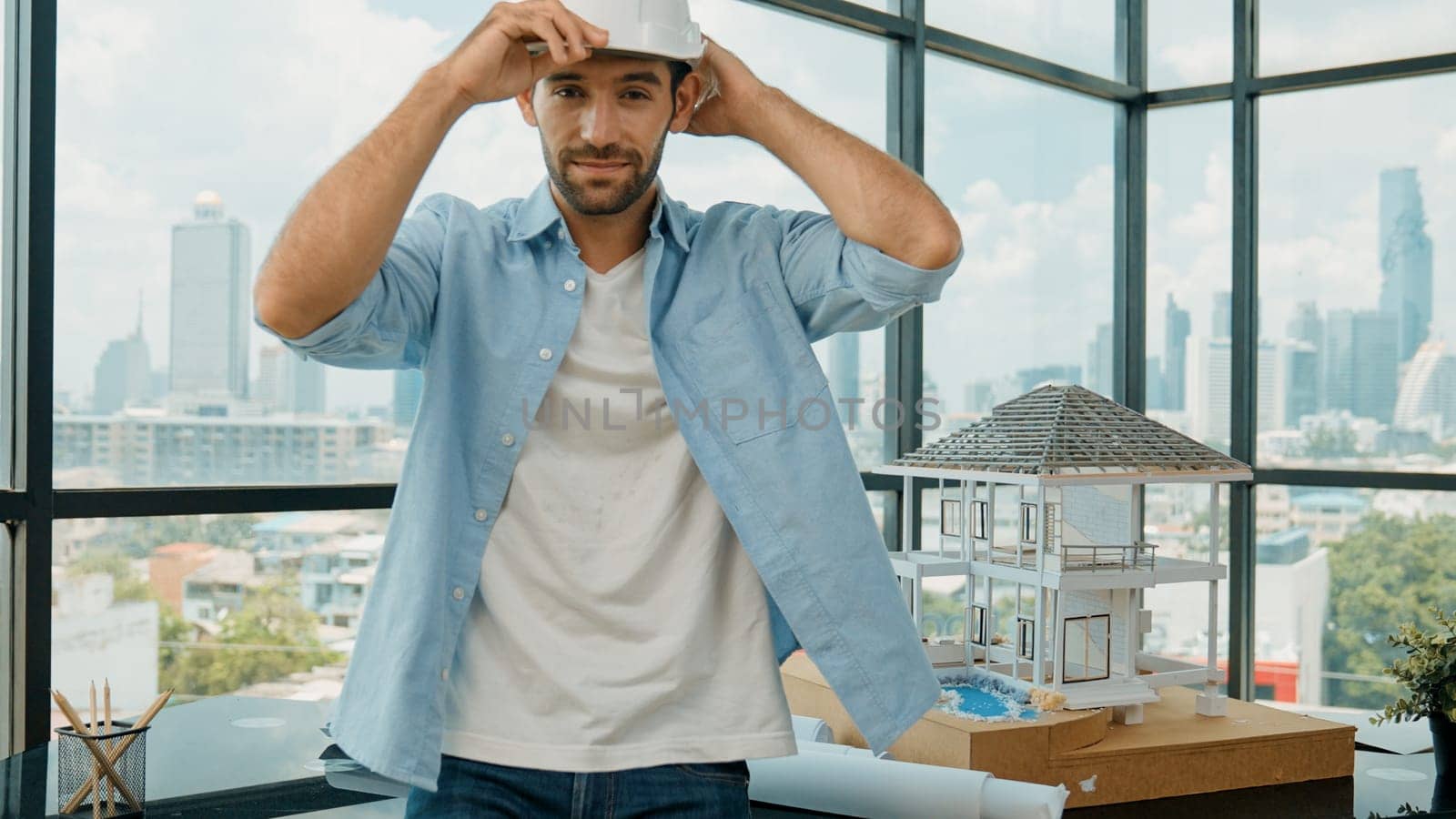 Portrait of smart architect engineer wear safety helmet while sitting on table with house model, project plan and architectural equipment surrounded by skyscraper view. Civil engineering. Tracery