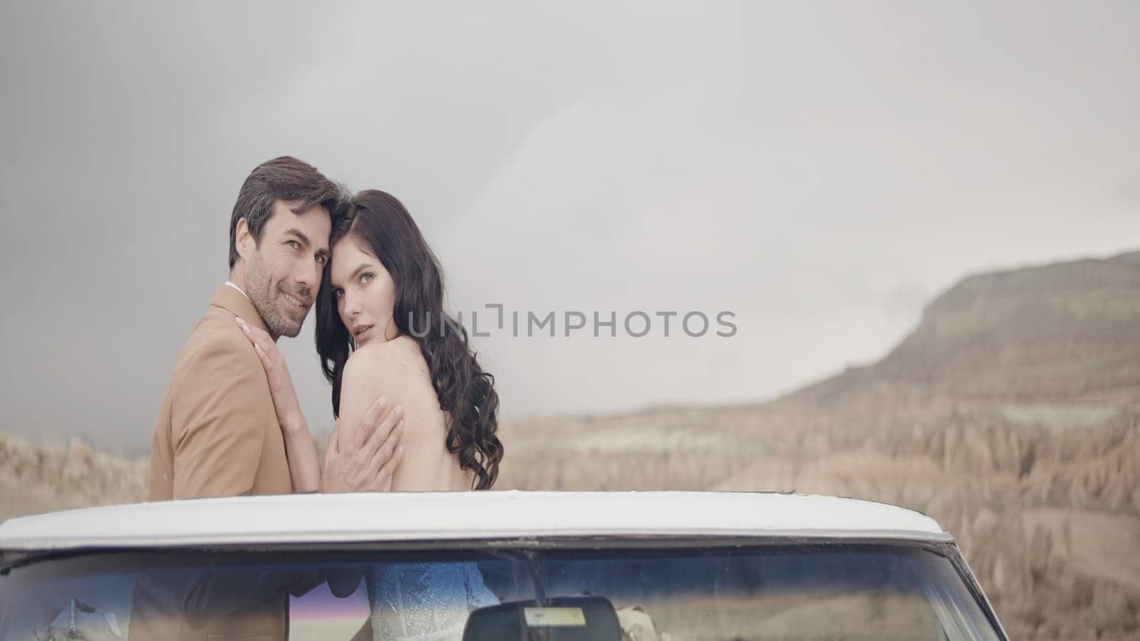Beautiful couple of newlyweds in convertible. Action. Elegant couple in love on convertible. Beautiful newlyweds are sitting on convertible on background of sunny sky and rocks by Mediawhalestock