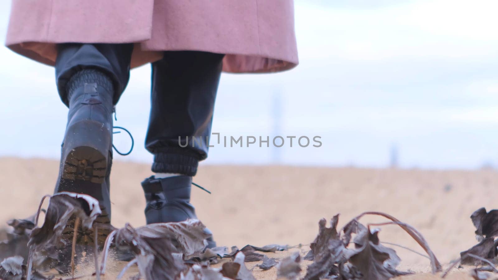 Rear view of female black shoes on the sand, close up view. Woman wearing black leather boots and pink coat walking away on sandy coast with dry tree leaves.