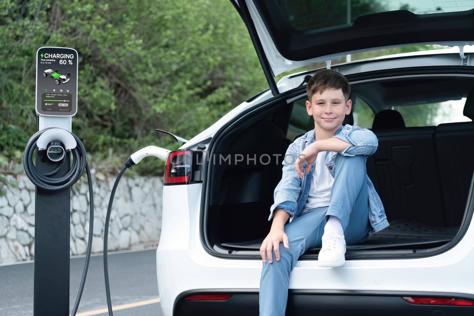 Little boy sitting on car trunk while recharging eco-friendly electric car from EV charging station. EV car road trip travel concept for alternative transportation powered sustainable energy.Perpetual