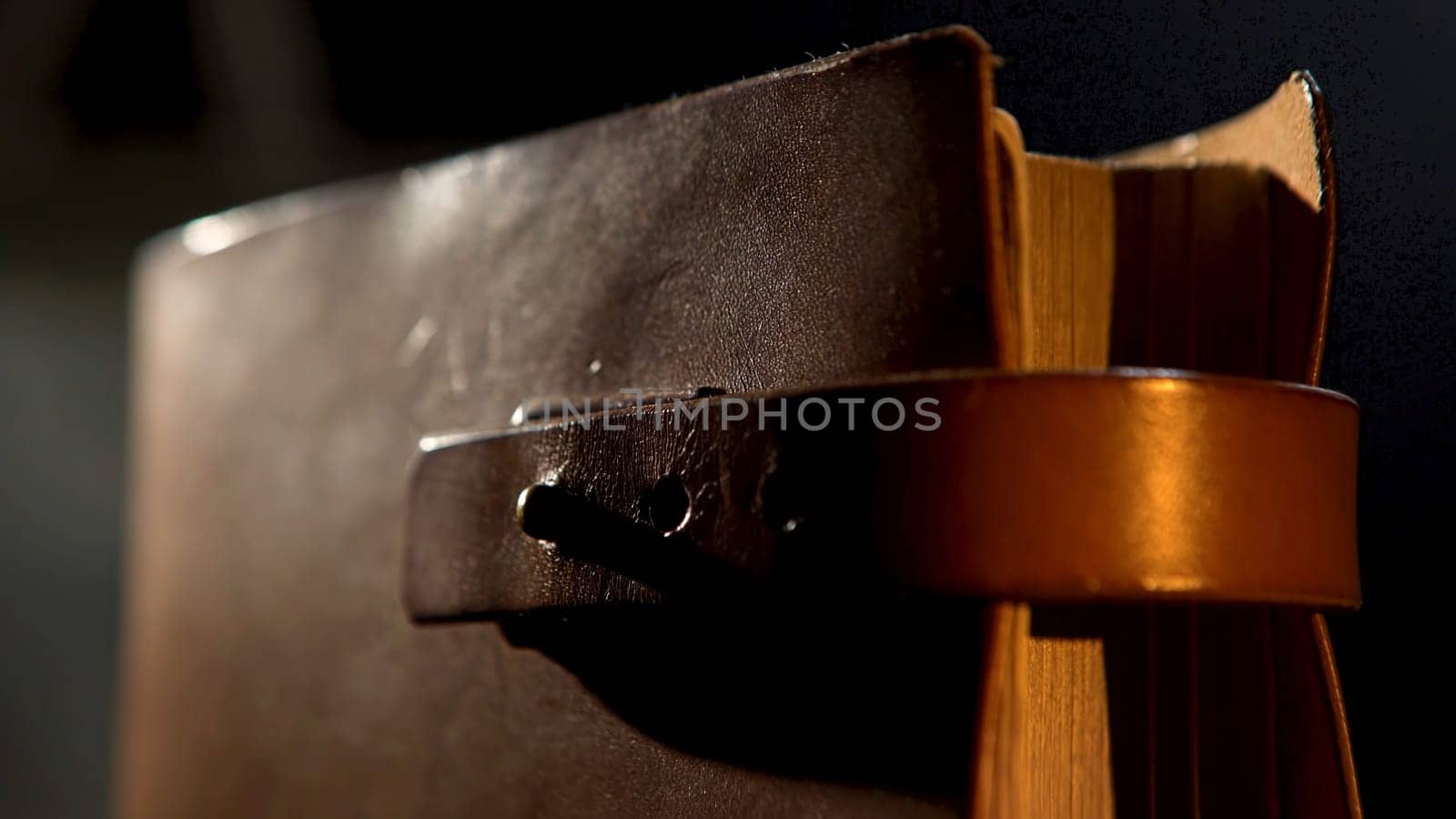 Vintage leather book with yellow pages. Close up of an ancient notebook with brown leather cover and a strap in vertical position isolated on black background under the dim light.