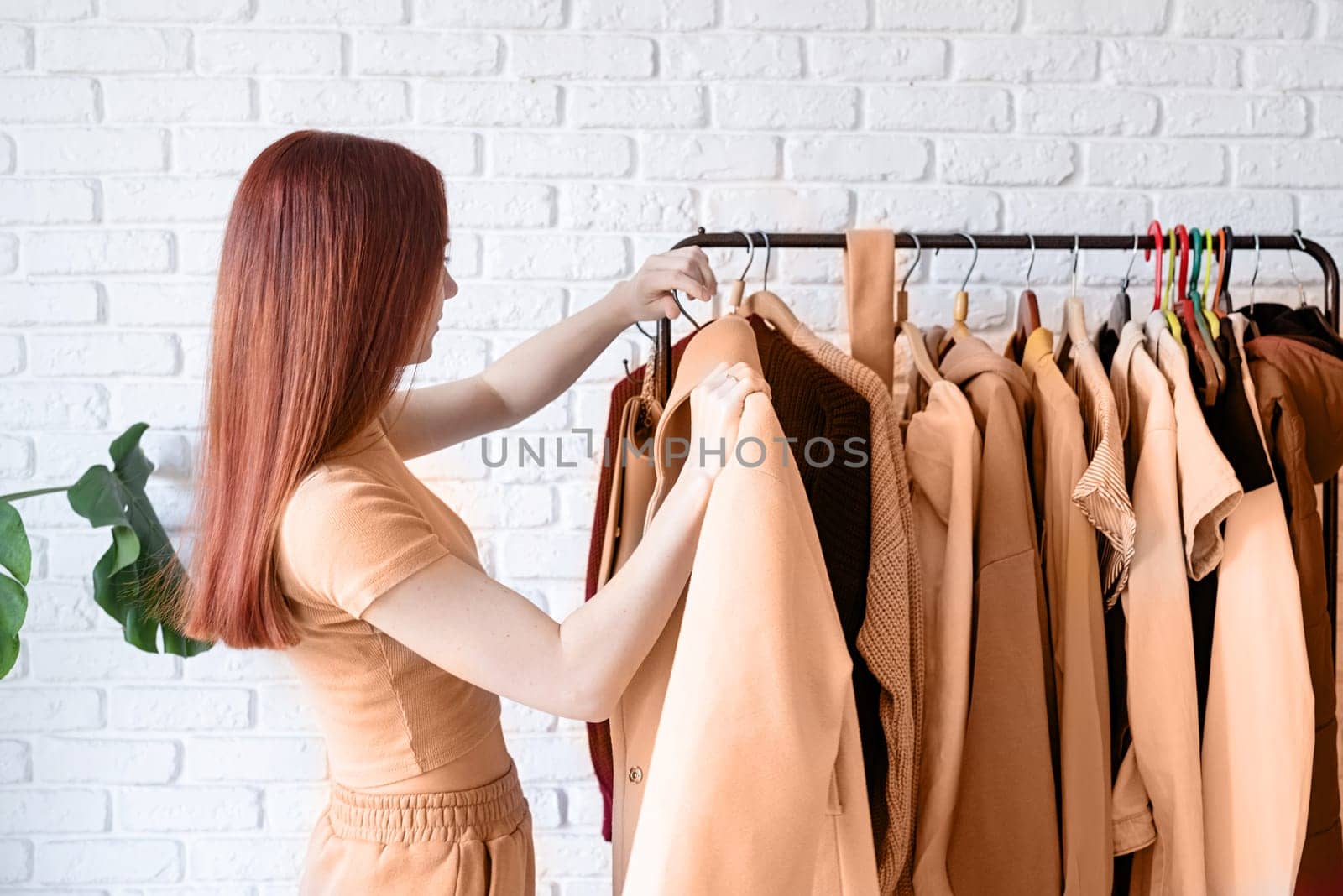 Color of the year 2024: Peach Fuzz. beautiful young woman in beige coat standing in front of hanger rack and choosing outfit dressing. Selection of a wardrobe, stylist, shopping. Clothes designing