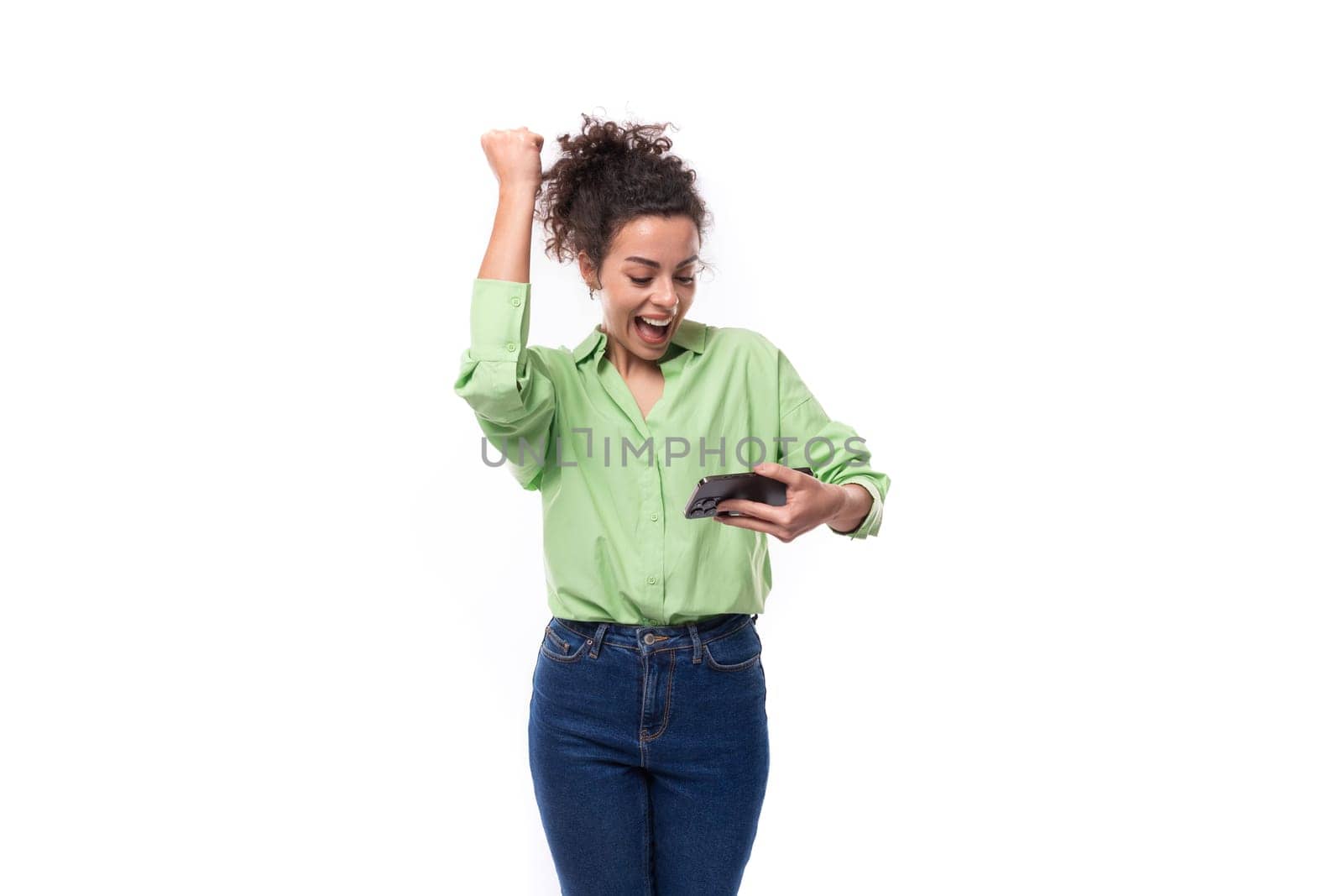 young charming brunette businesswoman with curly hair gathered in a ponytail rejoices looking into a mobile phone.