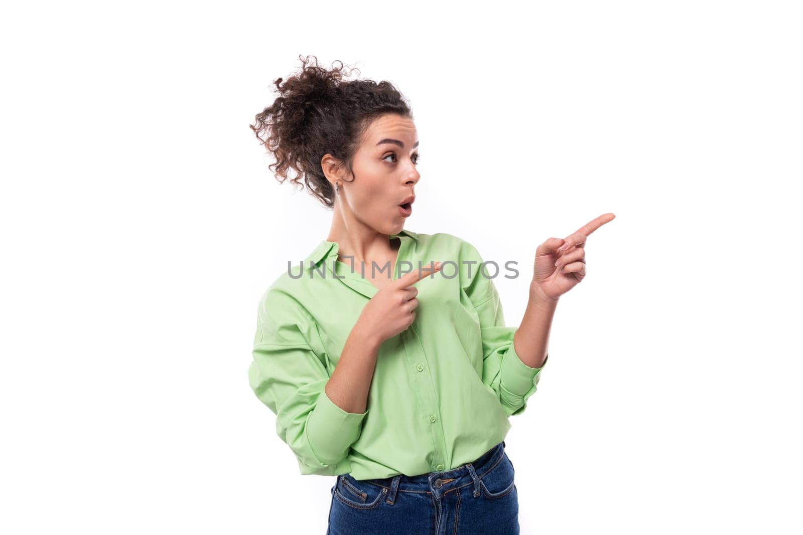 young brunette curly slender female model dressed in a light green shirt points with her hand at the space of the wall.