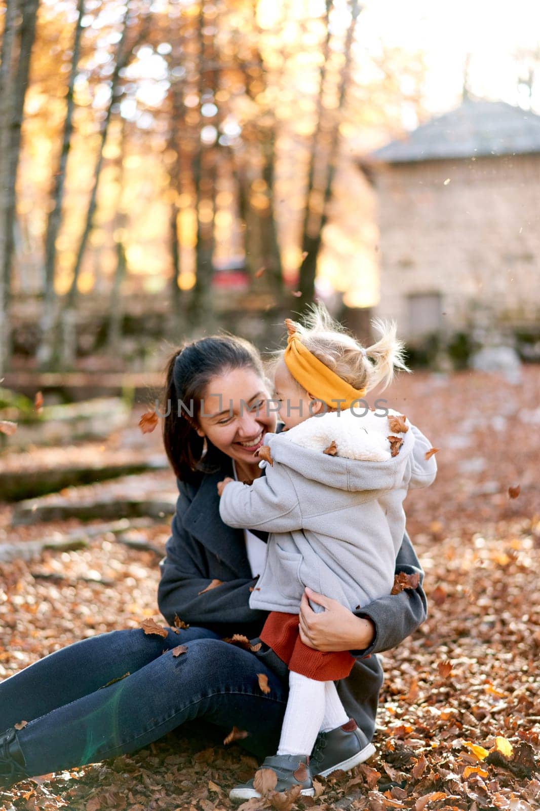 Little girl hugs her smiling mother sitting on the ground in the autumn forest under falling leaves. High quality photo