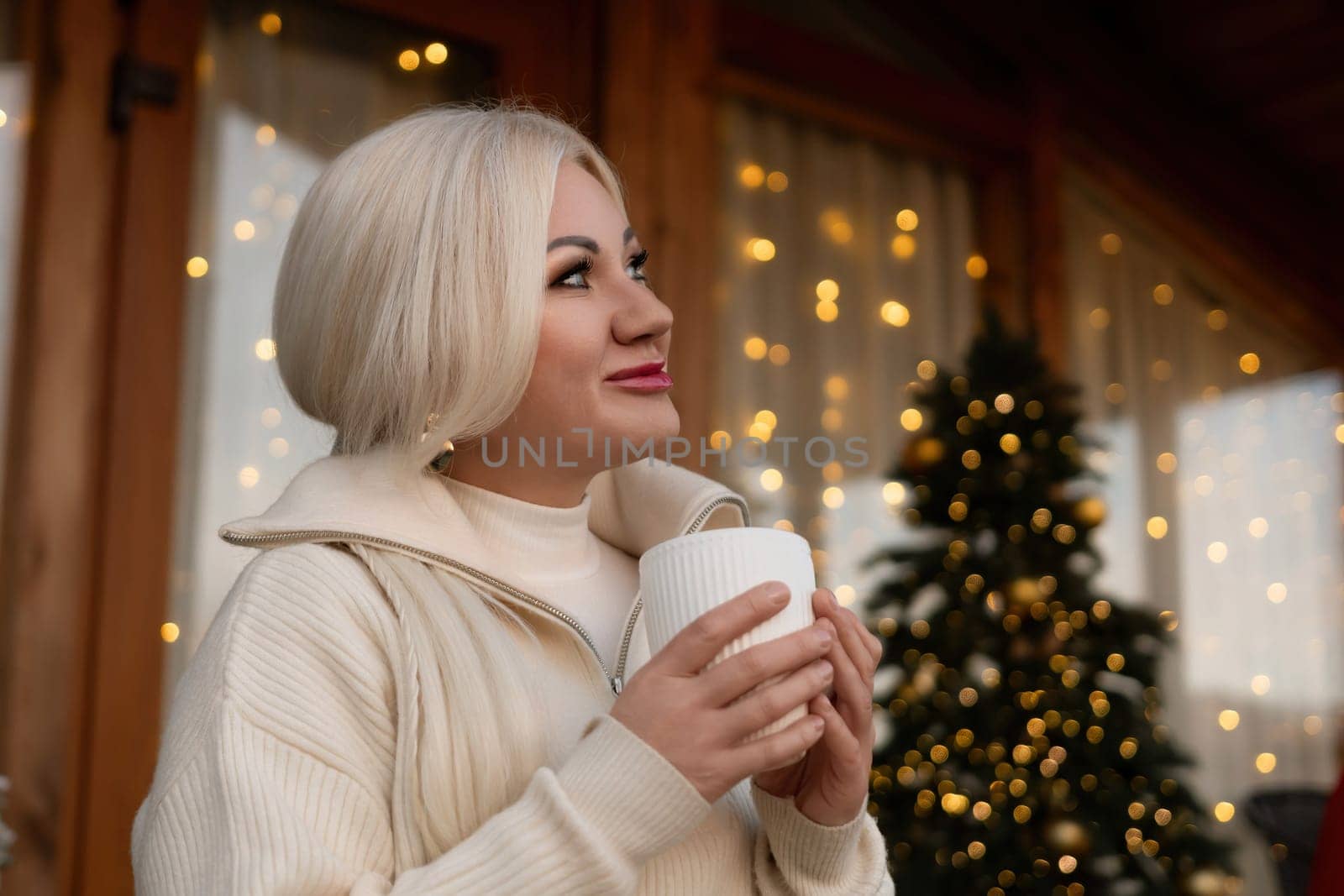 Happy blonde at home at Christmas time, drinking hot beverage from the white mug. Christmas tree in the background. Celebrating Christmas and New Year at home in cozy.