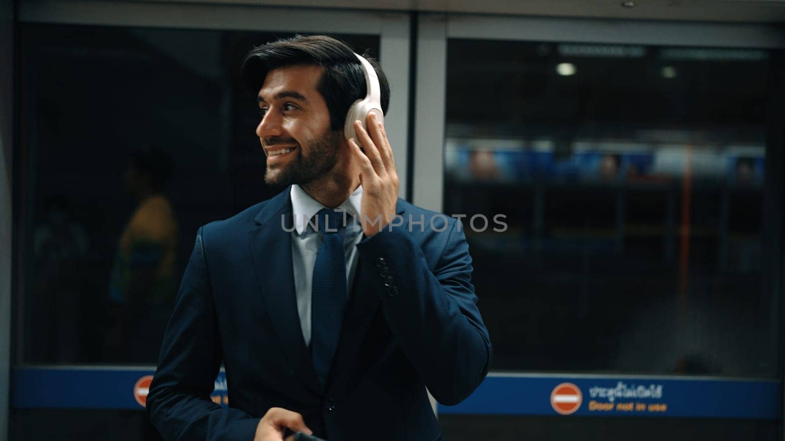 Closeup of smart executive manager listen relaxing music while waiting at train station. Happy male leader wear headphone and standing at door area. Public transport concept. Dark filter. Exultant.