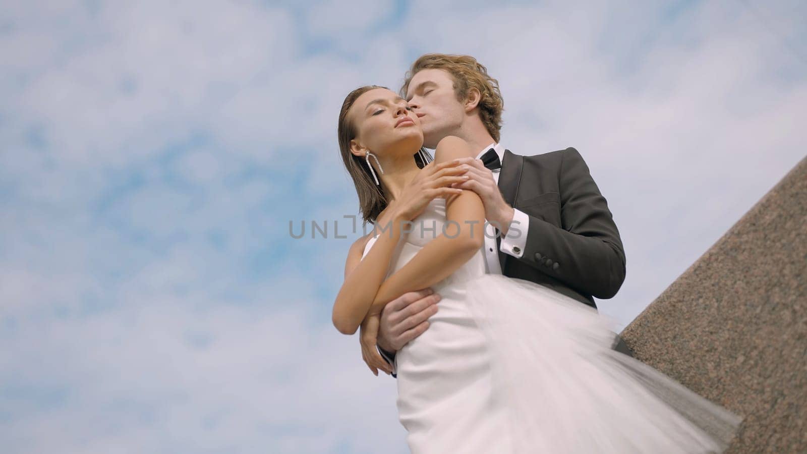 Bottom view of stylish caring bride and groom on blue sky background with clouds. Action. Man in black and white suit kissing woman cheek in white dress