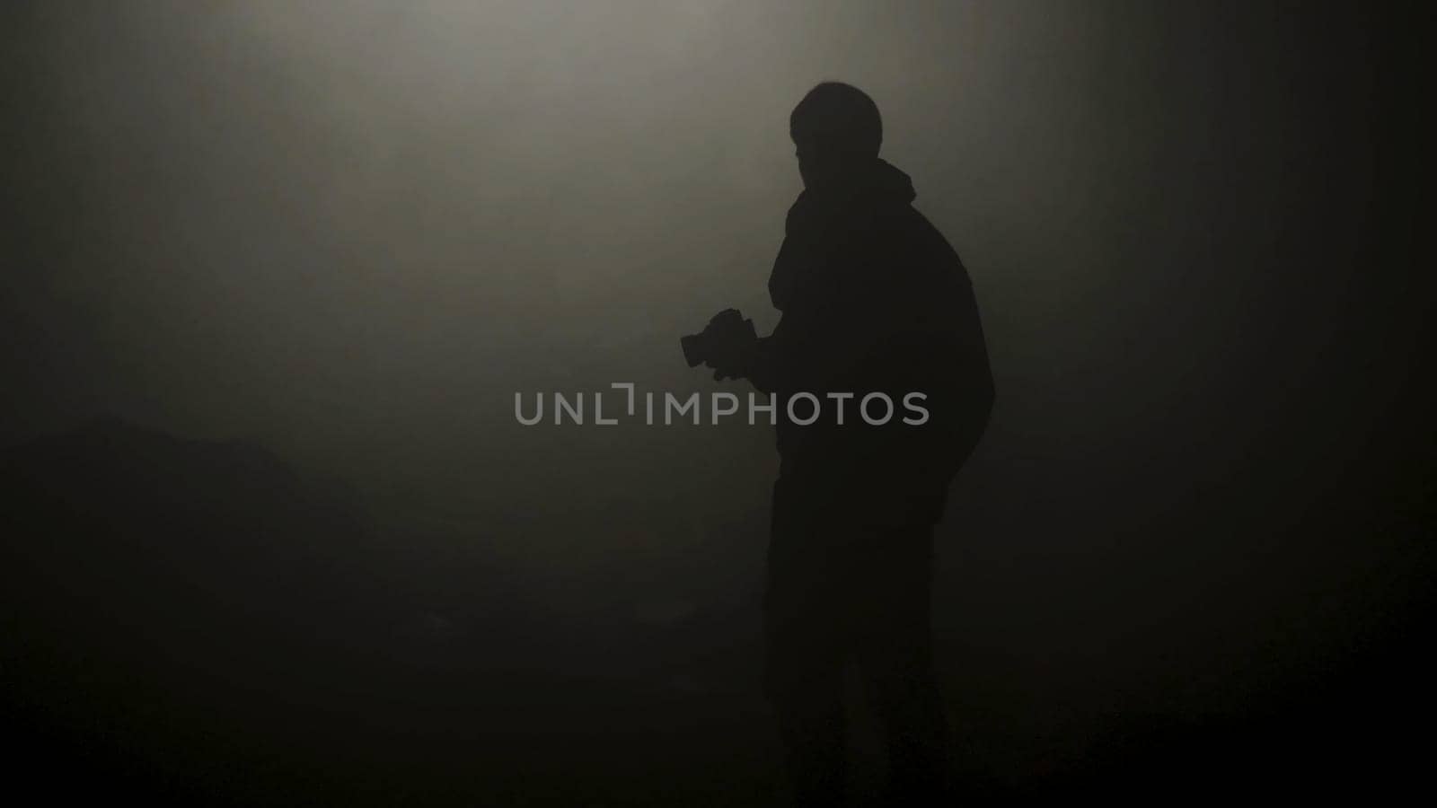 Black silhouette of man standing in smoky dark. Stock footage. Mysterious silhouette of young man stands in darkness lit only by dim light in haze.