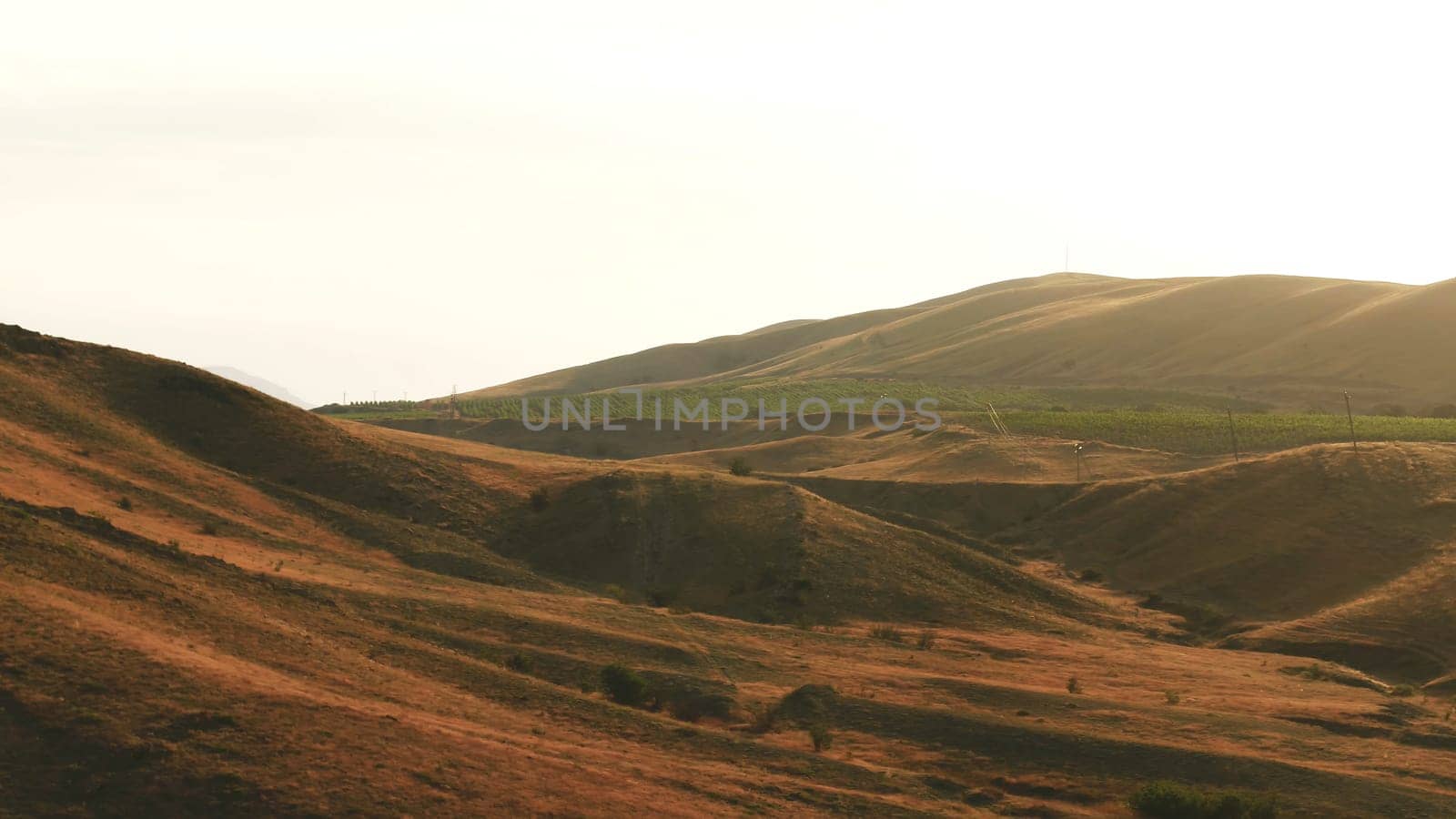 Aerial view of exciting red hills covered with shrubs and large green farmlands against the evening sky in summer day. Amazing colorful landscape
