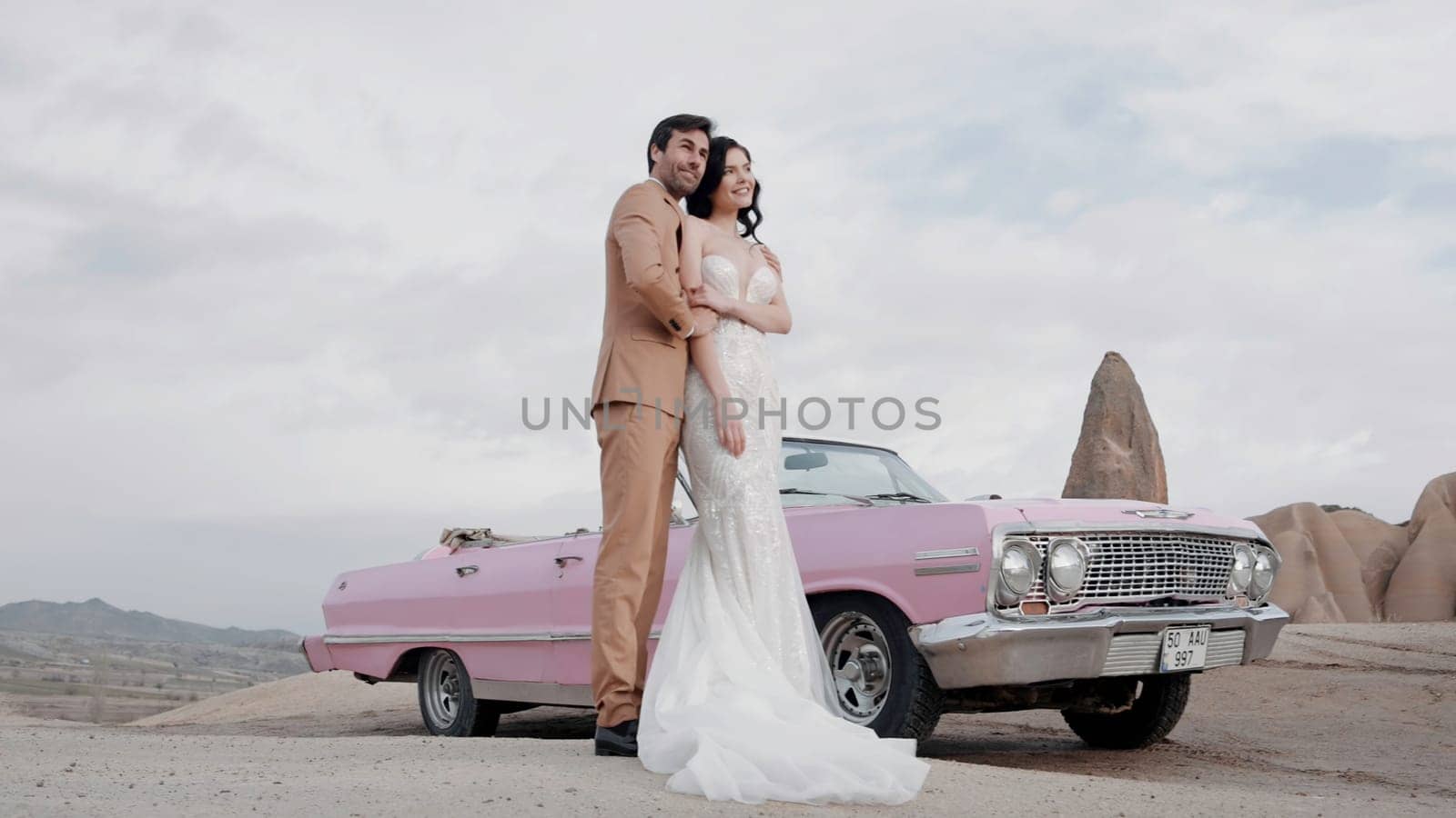 Elegant wedding photo shoot. Action. A beautiful bride in a wedding dress with a groom in a brown suit posing for a photographer on camera with happy faces in the desert next to a pink small car.
