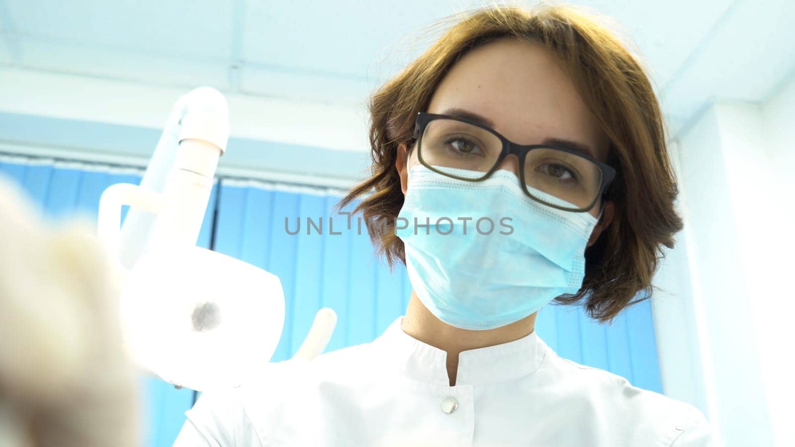 First person view of a young female dentist in mask approaches with Tools. Media. Young doctor standing over the patient, looking at camera. by Mediawhalestock