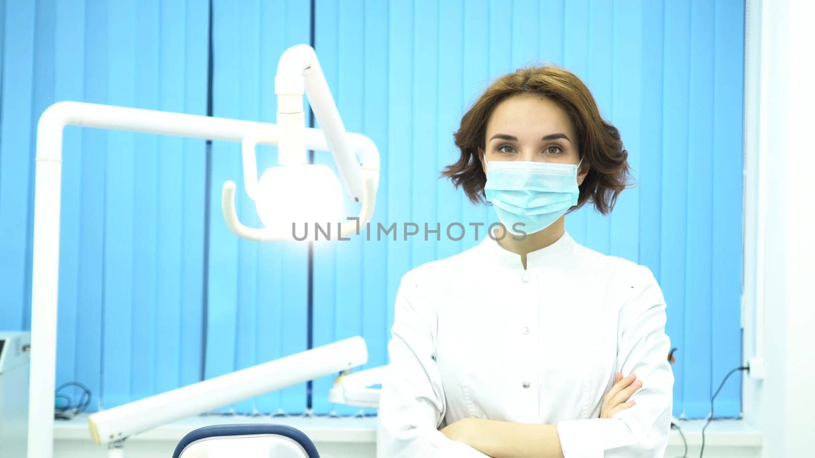 Portrait of a dentist putting on the mask looking at camera at the dental clinic. Female dentist assistant standing arms crossed with surgical mask on her face