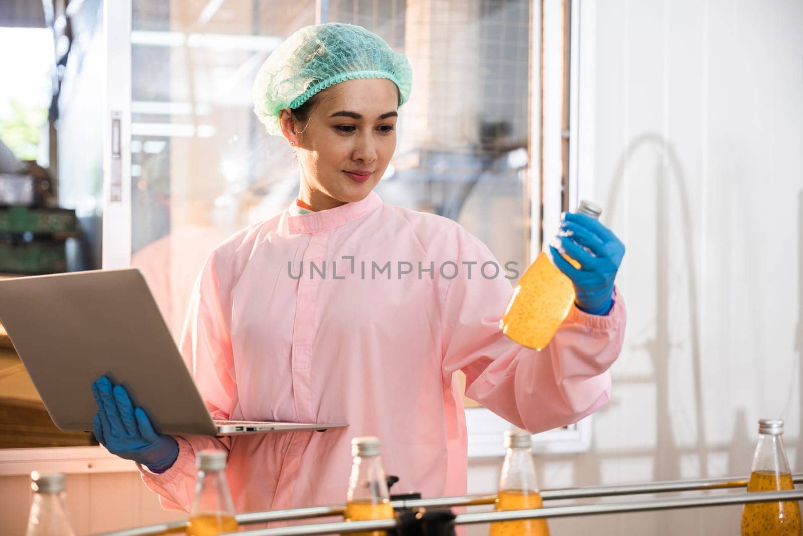 Engineer woman monitors bottles on conveyor belt using laptop in beverage factory. Quality control manager guarantees drink's excellence. Technology-driven liquid control is the answer.