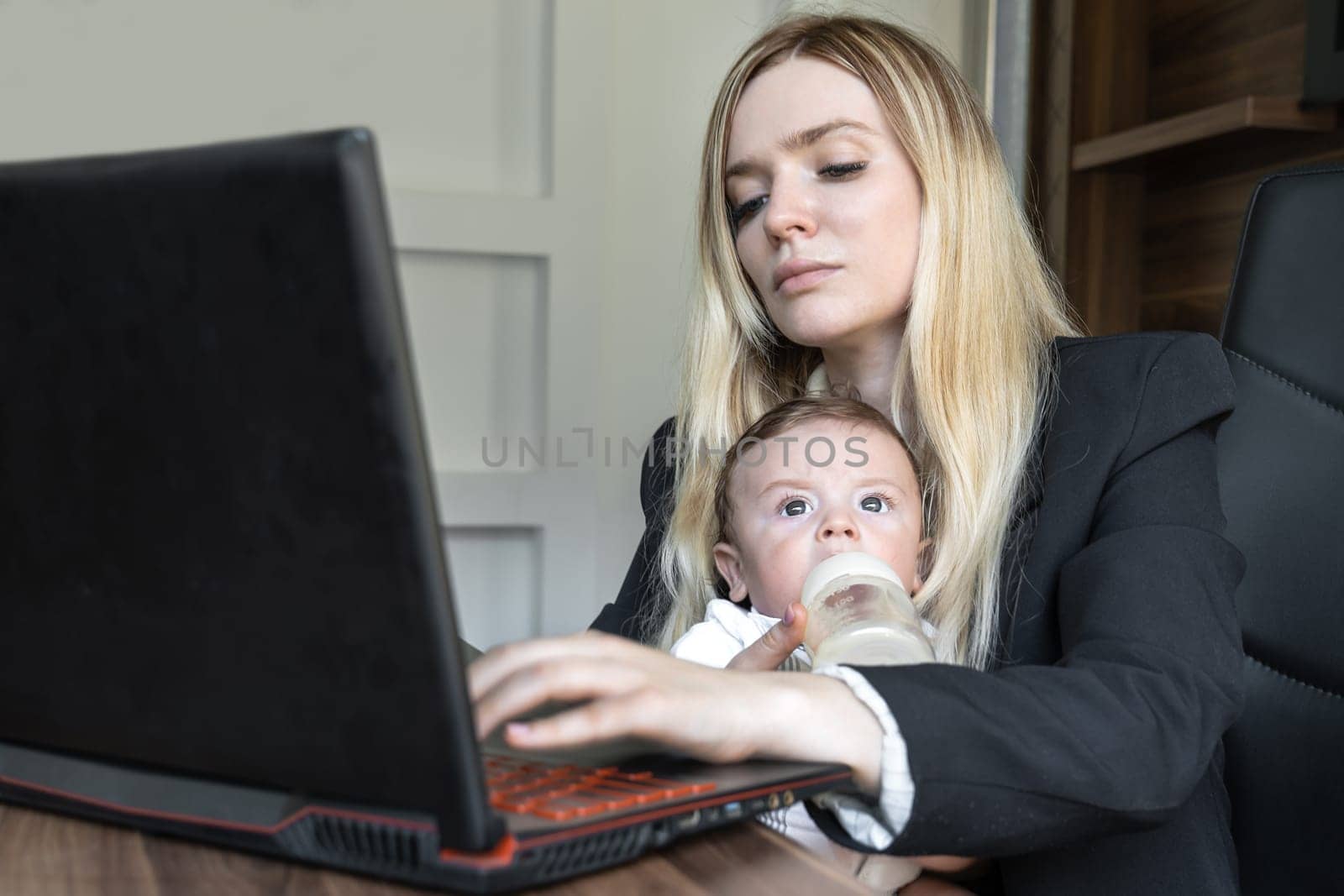 Concept of combining business and caring for a newborn child, young business woman feeds a baby from bottle in office, combining it with work on laptop.