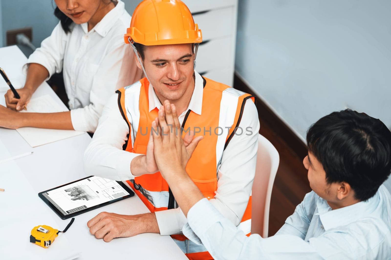 Diverse group of civil engineer and client celebrate and high five after make successful agreement on architectural project, reviewing construction plan and building blueprint at meeting table.Prudent