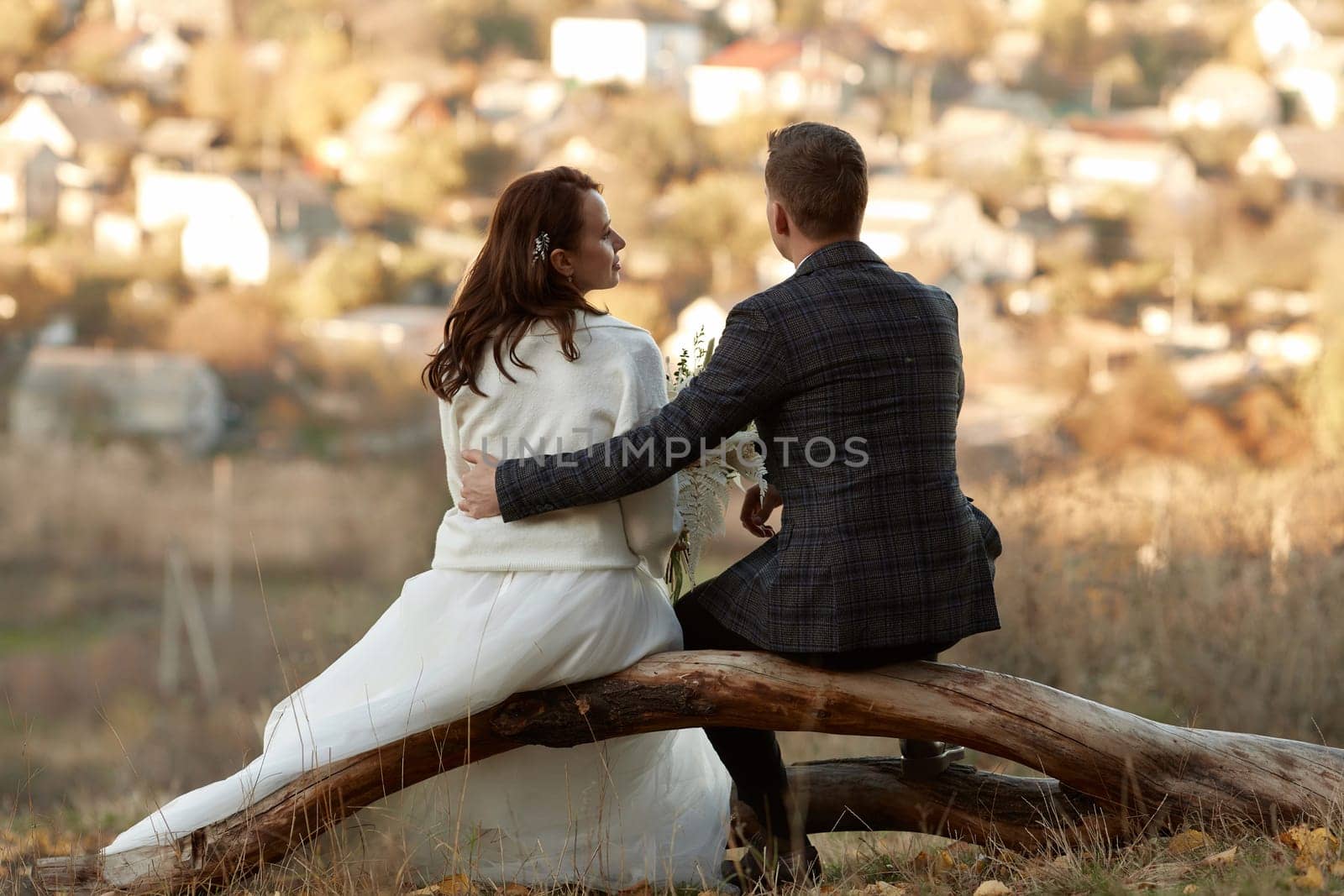 bride in white wedding dress and groom are sitting on tree in nature, back view