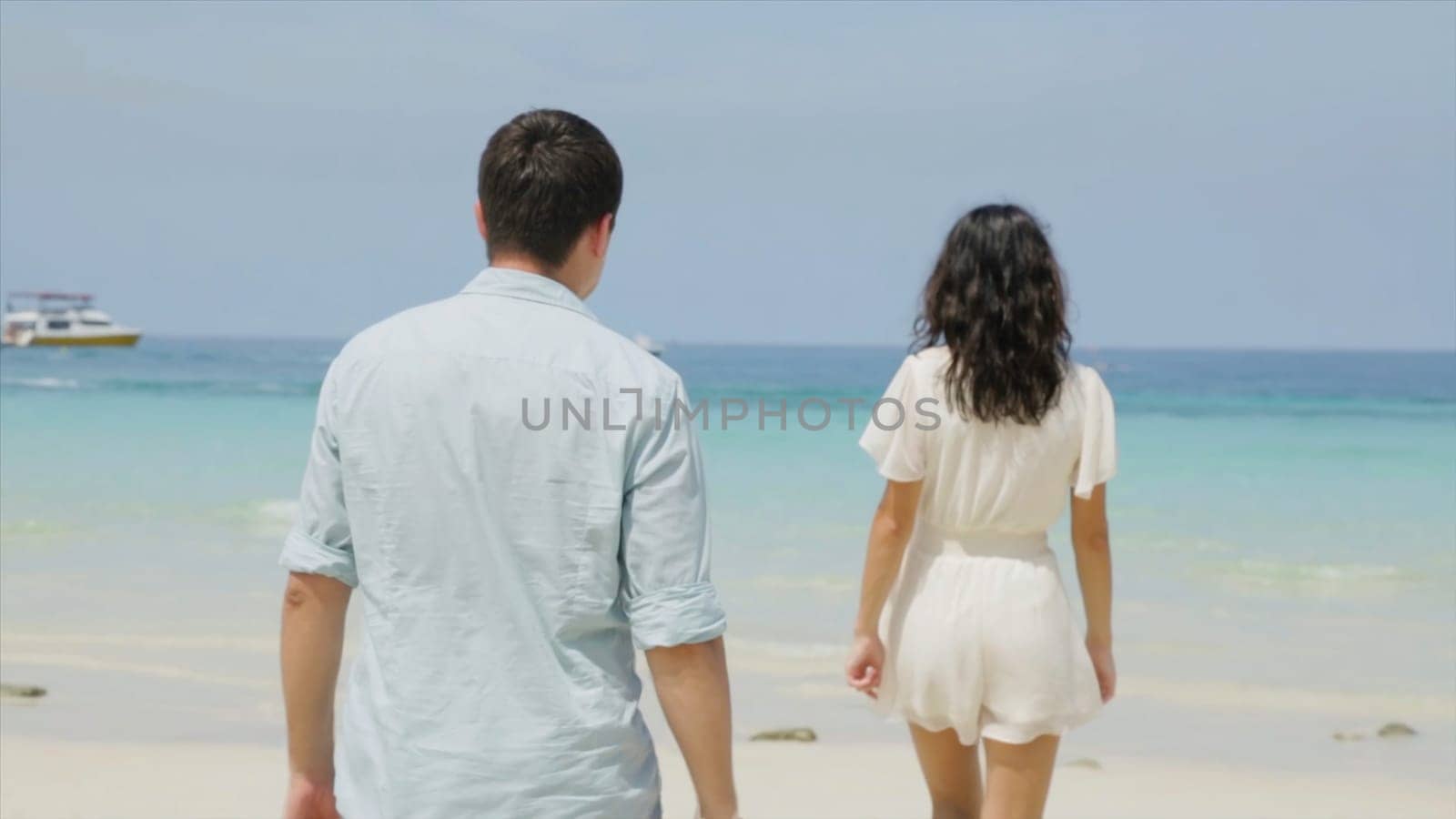 Cute couple in white clothing goes hand beach. Couple of teenagers running and flirting on the beach shore near the water. Woman walking on romantic honeymoon beach holidays holding hand of boyfriend following her, view from behind. A young girl holding hands on the beach.