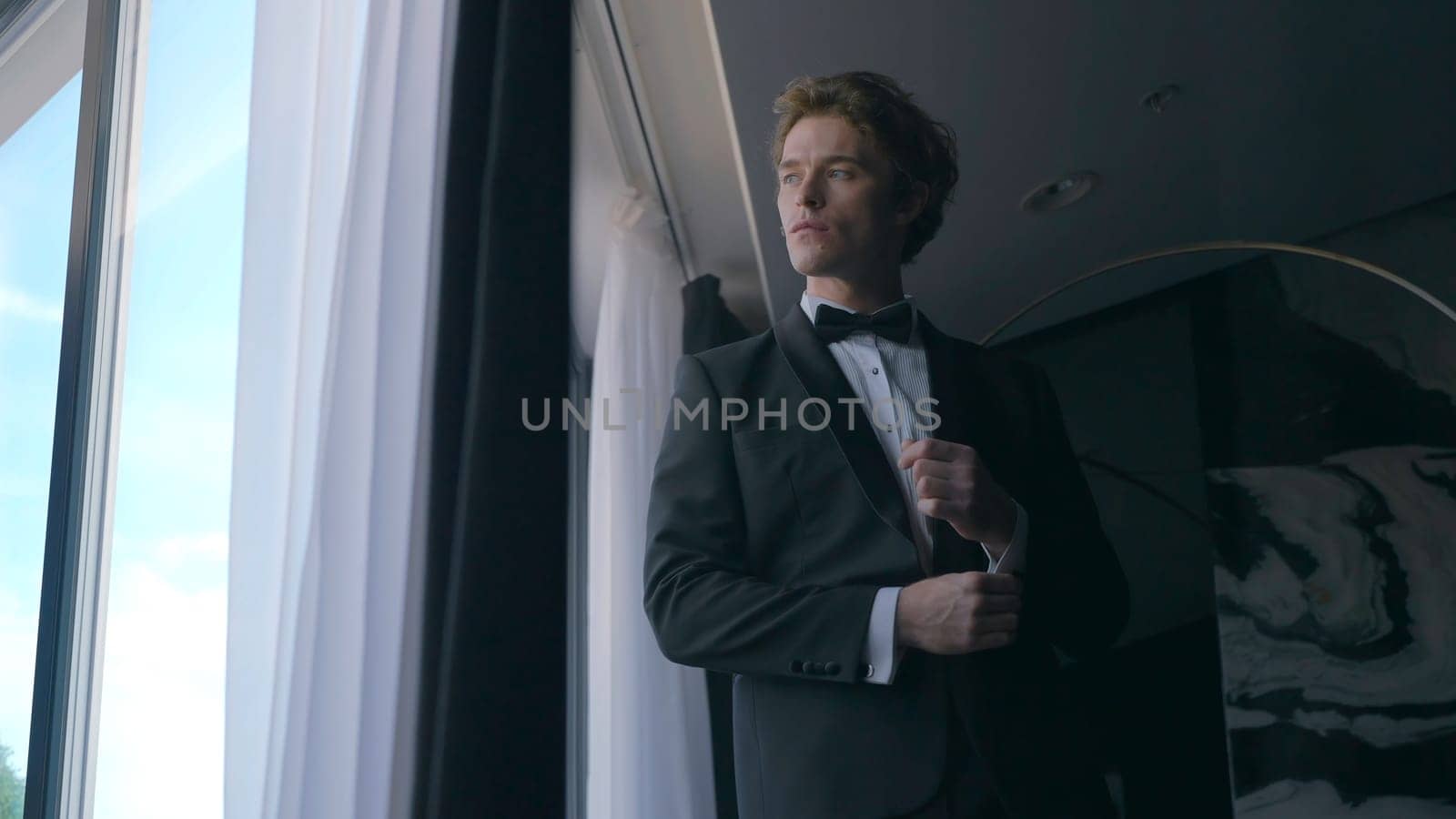 Handsome man in suit by window. Action. Gentleman in suit with bow tie looks out window. Attractive man in suit is preparing for celebration.