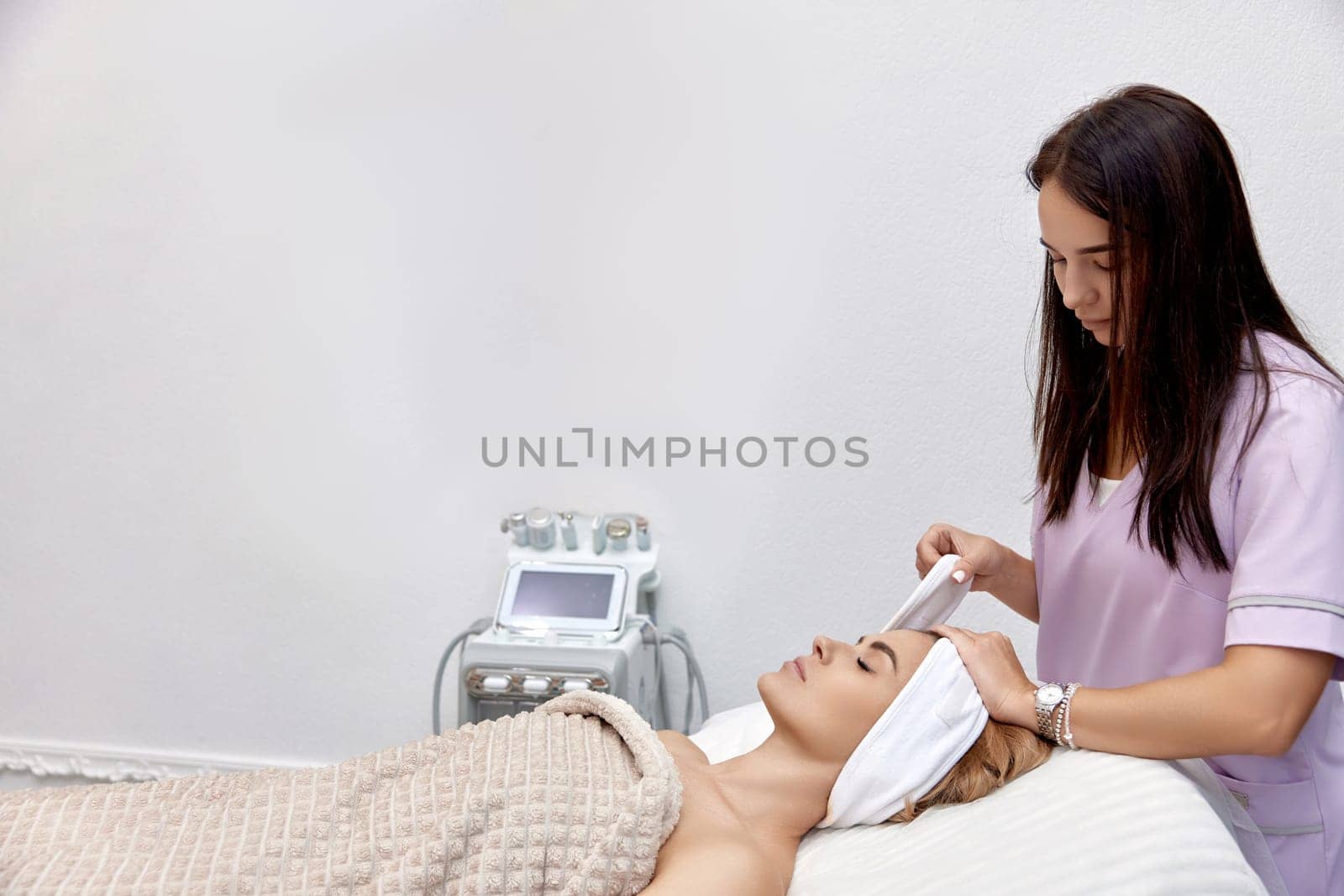 Cosmetologist putting white headband towel on female patient head in beauty salon