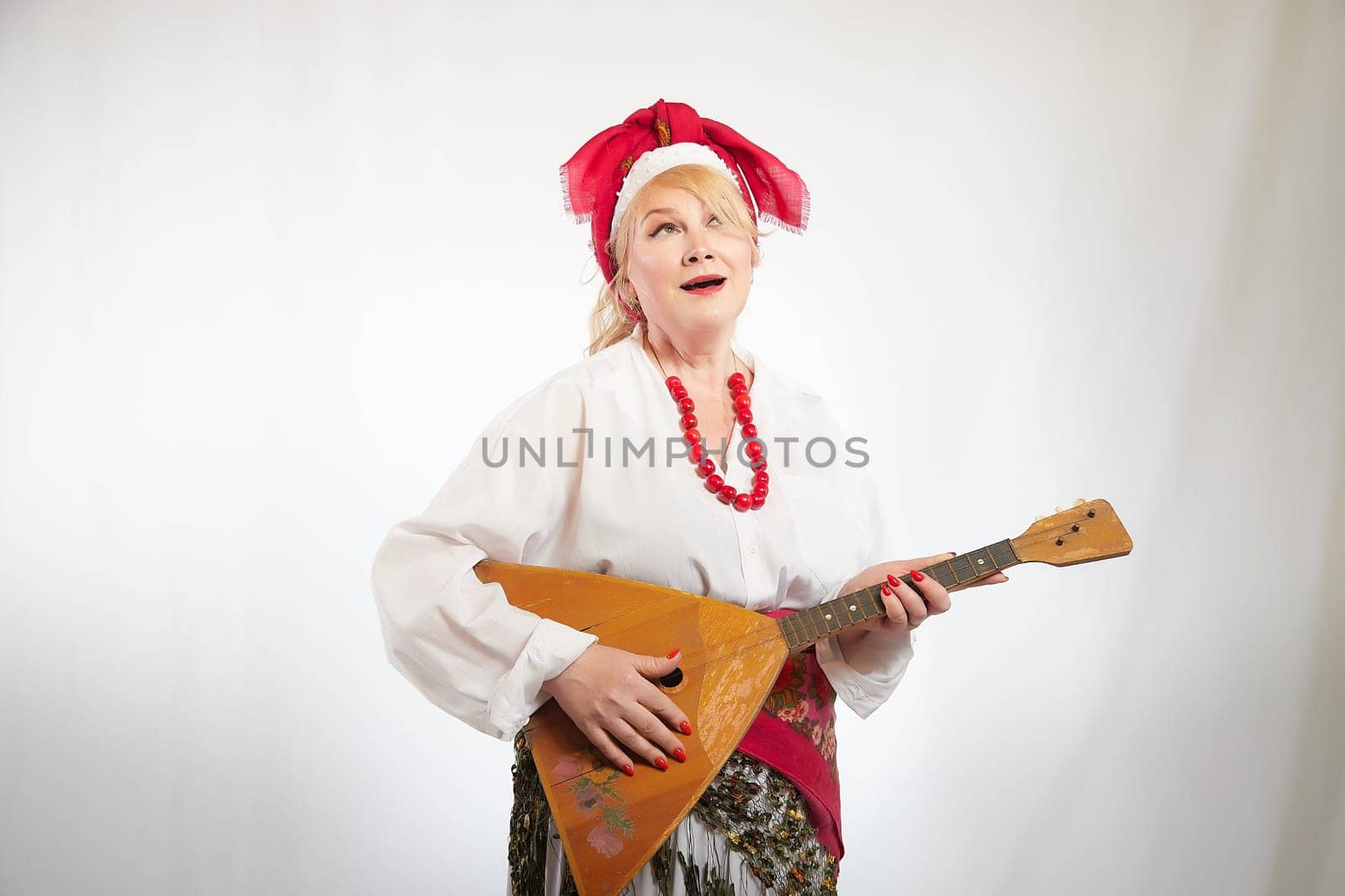 Cheerful funny adult mature woman solokha with musical balalaika. Female model in clothes of national ethnic Slavic style. Stylized Ukrainian, Belarusian or Russian woman poses in a comic photo shoot by keleny