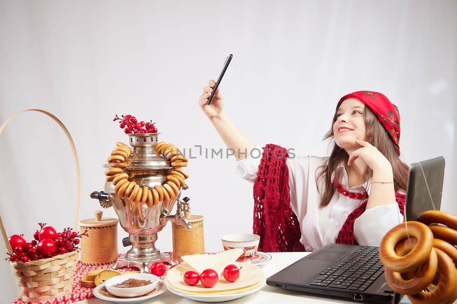 A fashionable modern girl in stylized folk clothes at a table with a samovar, bagels and tea takes a selfie on the Orthodox holiday of Maslenitsa and Easter. Funny photo shoot for a young woman by keleny