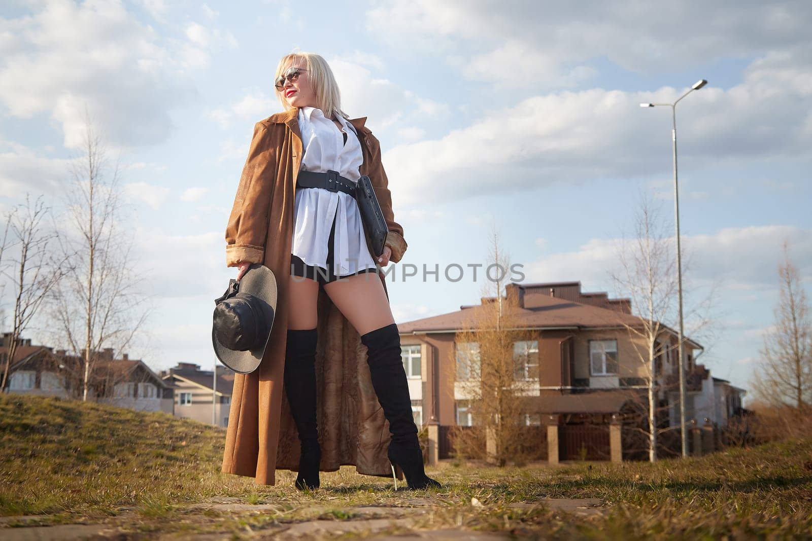 stylish, fashionable, imposing blonde girl in black glasses, a white shirt, a short skirt and a long raincoat with a laptop and hat on the street of the town. Defiantly dressed confident businesswoman by keleny