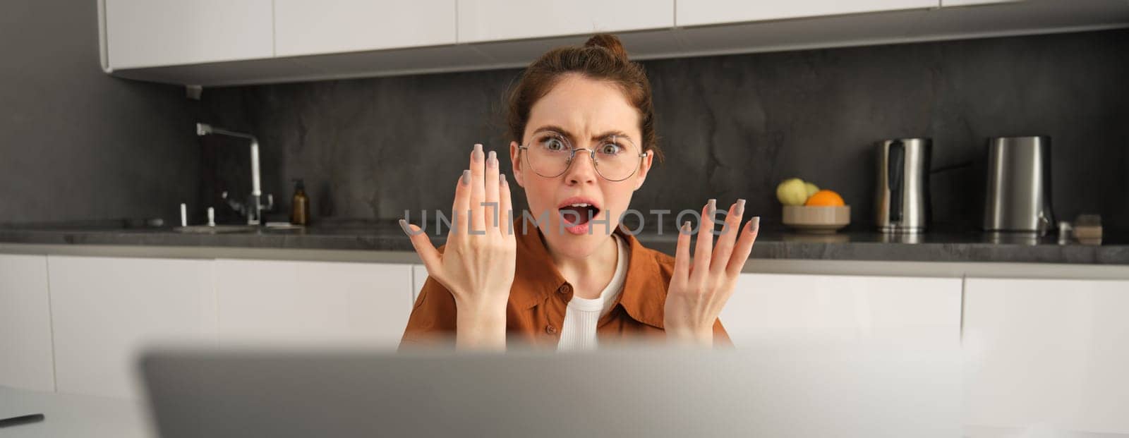 Portrait of woman has disappointed reaction, seeing upsetting news on laptop screen, looking frustrated or shocked, sitting at home in kitchen.