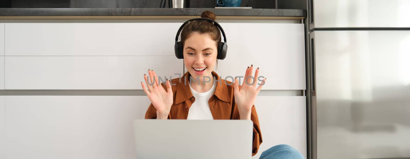 Portrait of happy young woman, freelancer working from home, remote worker sits on floor with laptop and headphones, looks surprised and excited at screen.