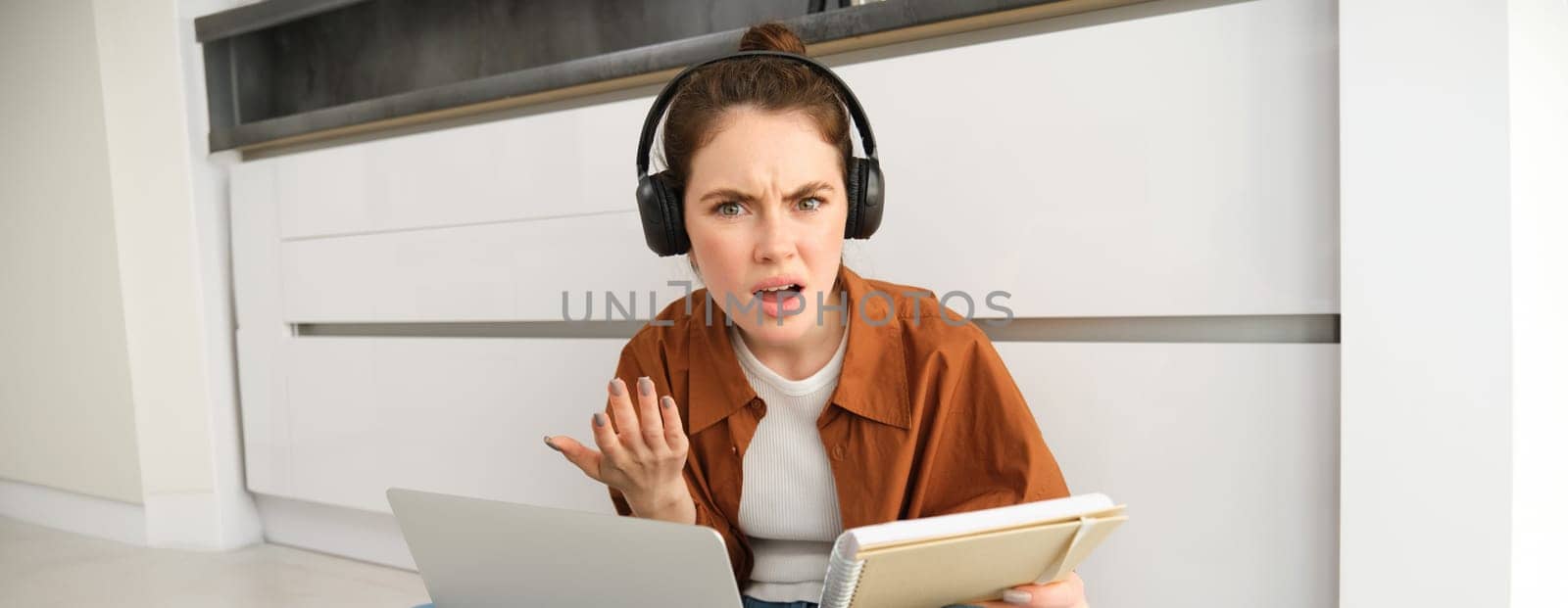 Portrait of annoyed and confused woman in headphones, sits on floor with laptop and notebook, looks frustrated, has issues in doing homework or studying.
