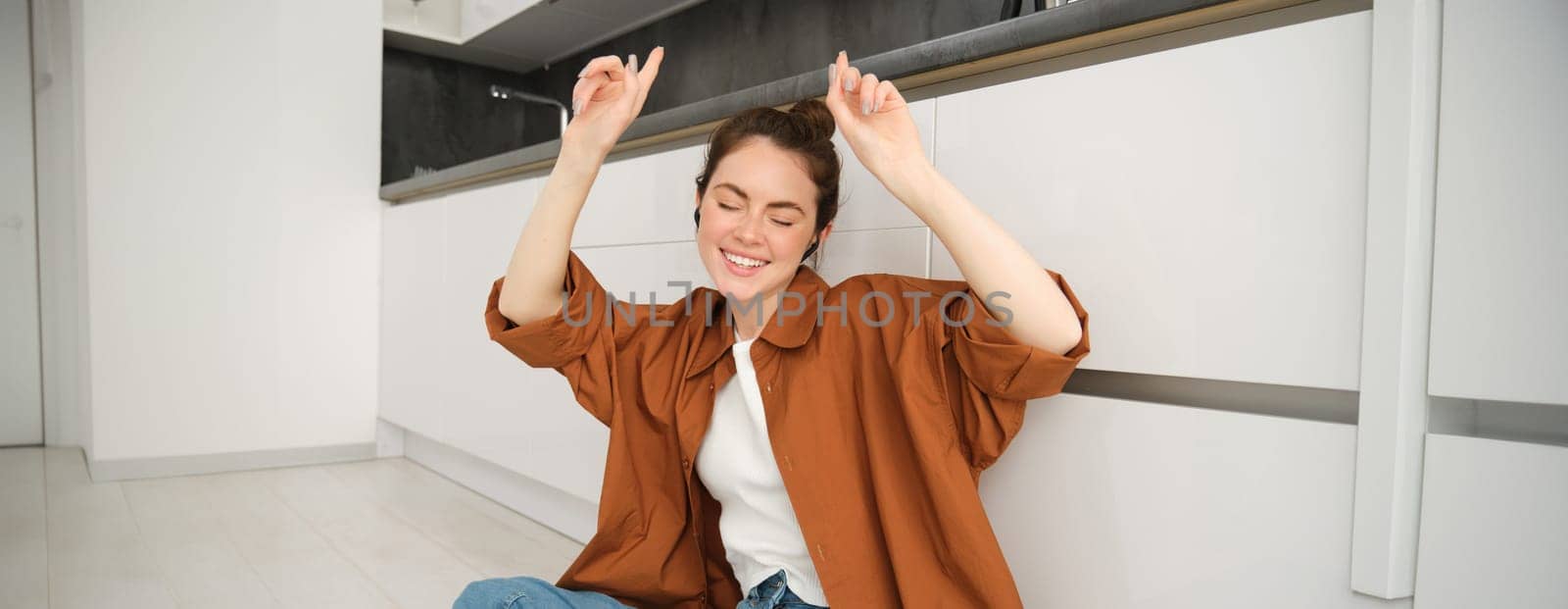 Dancing girl sits on kitchen floor, moving to the rhythm, enjoying sound quality in wireless headphones, listening to music in earphones.