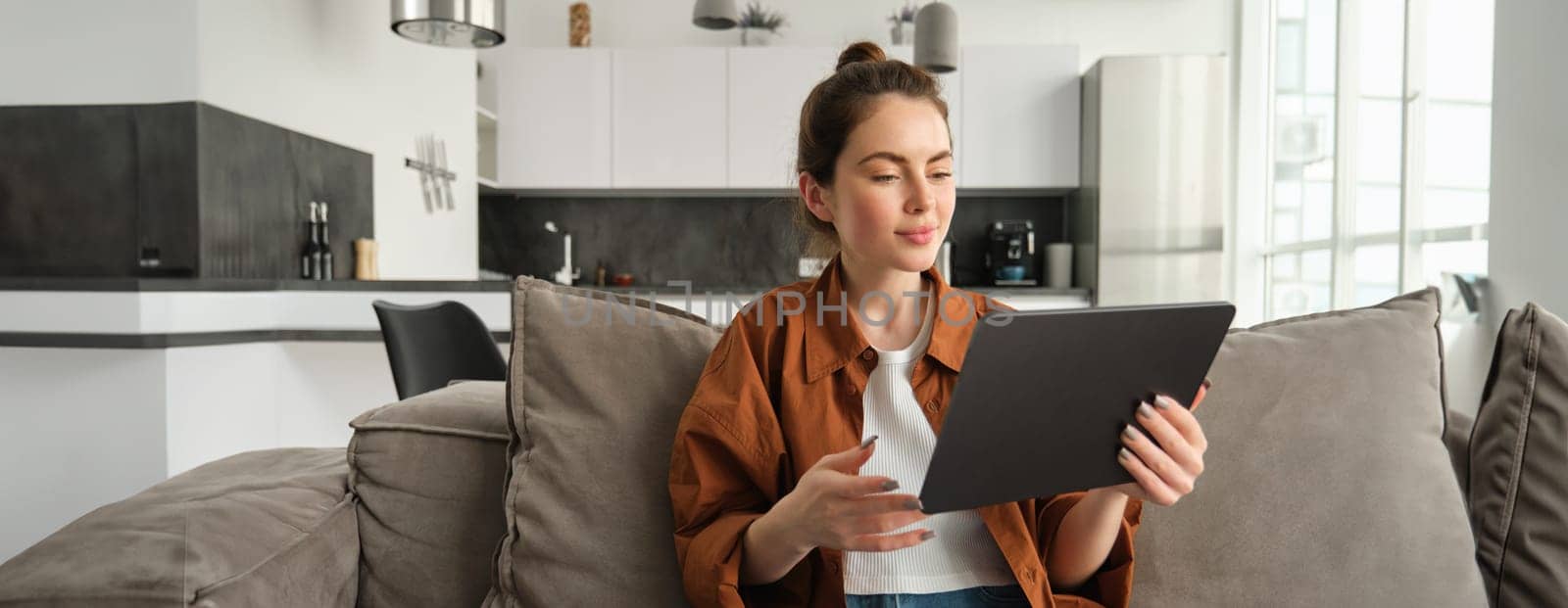 Portrait of female model reading on digital tablet e-book, watching tv series on her gadget application, sitting in living room at home.