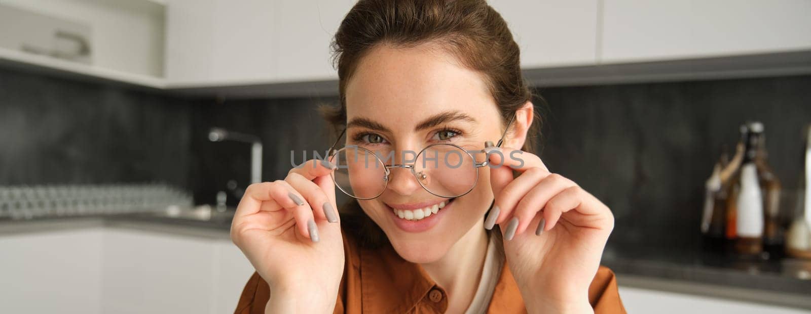 Close up portrait of young modern woman, wearing her glasses for reading, sitting at home in kitchen, smiling and looking at camera.