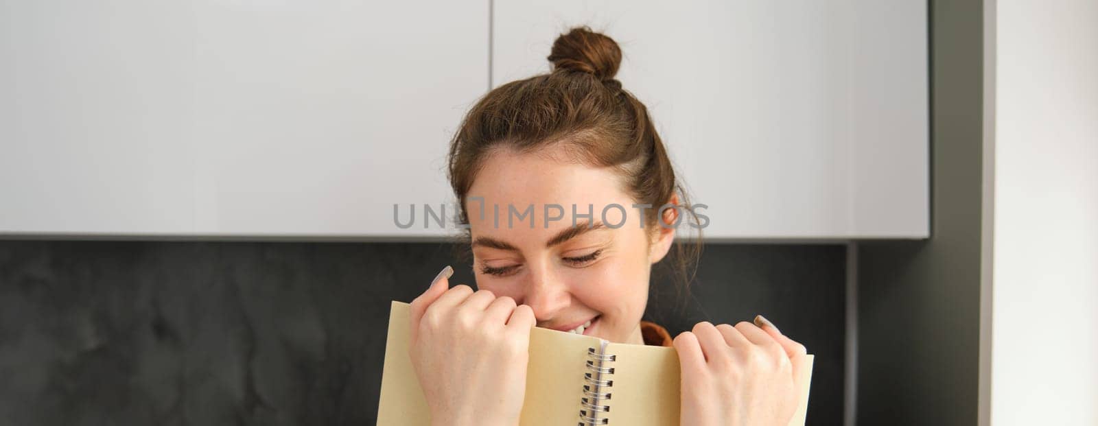 Coquettish young woman smiling, hiding face behind notebook, laughing and smiling, standing in the kitchen.