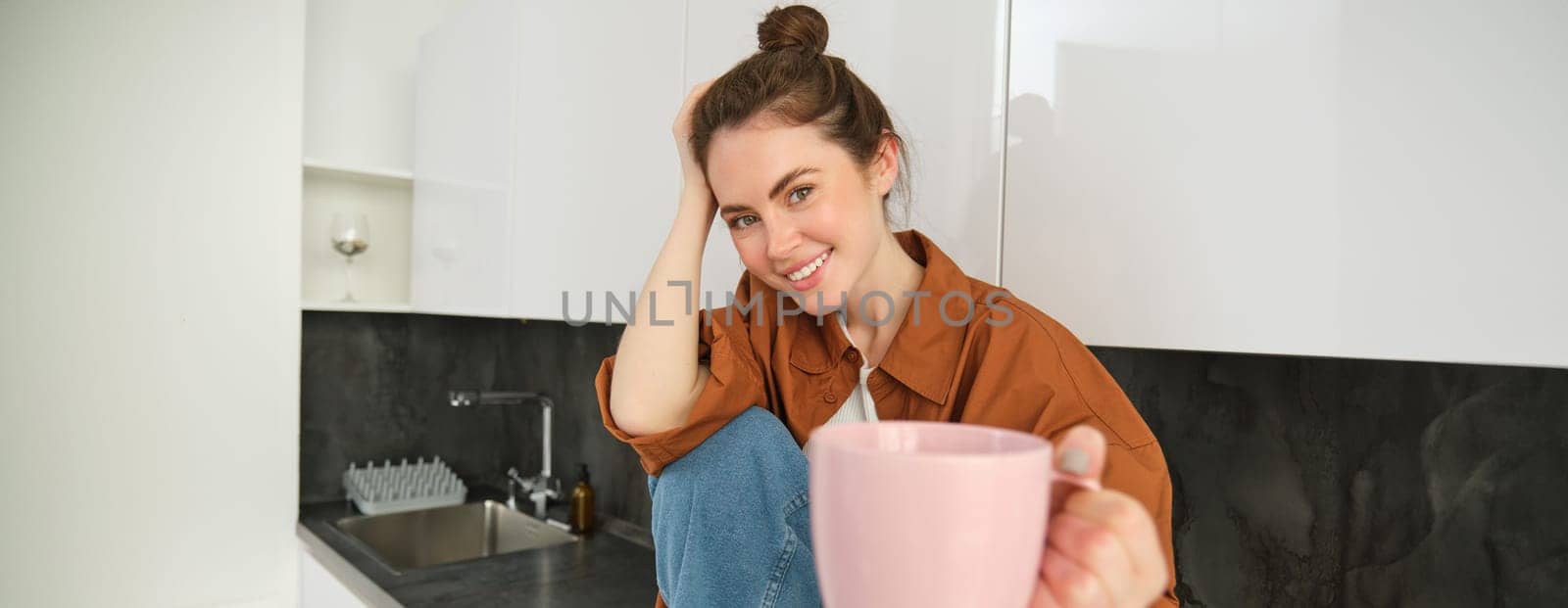 Pretty young smiling woman sits in kitchen, offers you cup of coffee, extends hand with pink mug, sharing her drink with someone.