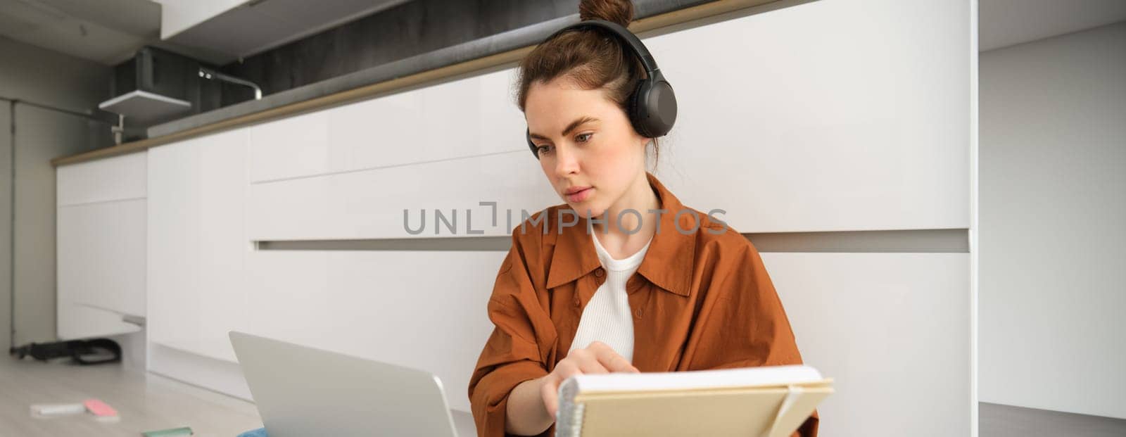 Serious-looking girl student studying at home, sits with laptop and notebook on floor, doing homework project on computer, has online course lesson, wears headphones.