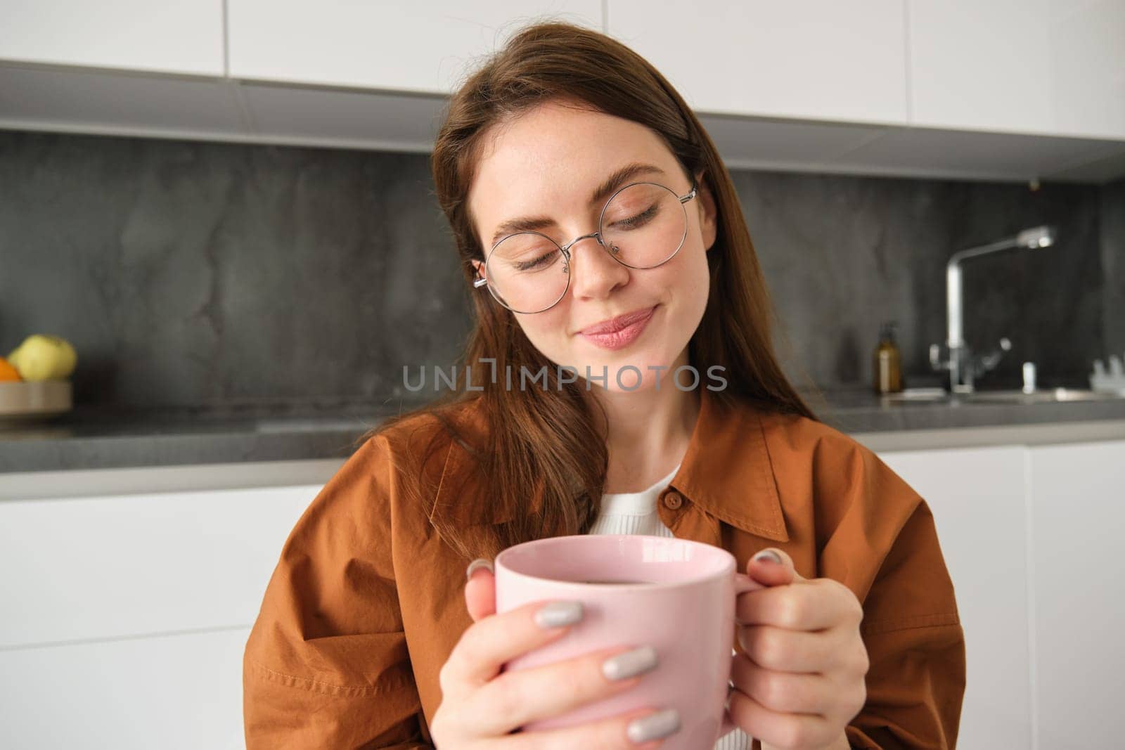 Portrait of cute, lovely young woman in glasses, sitting in kitchen, holding mug, drinking delicious tea.