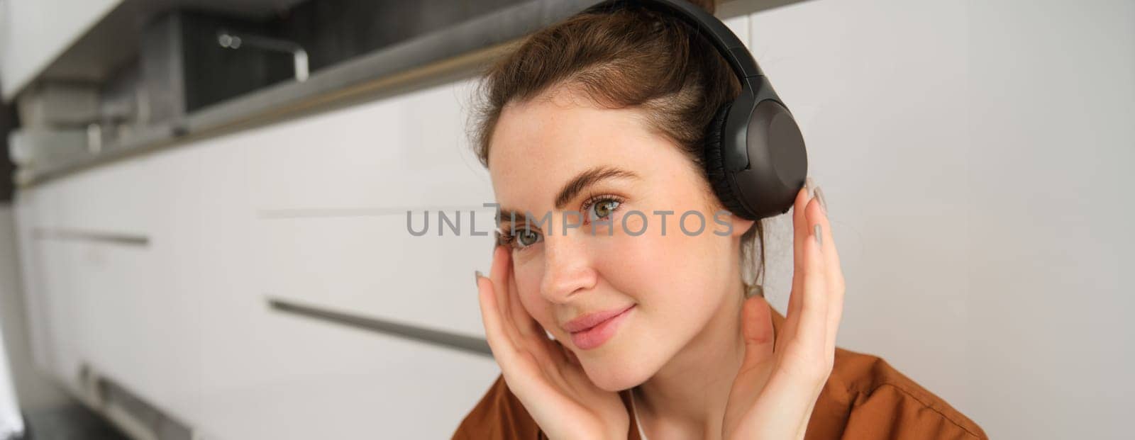 Image of modern woman, beautiful girl enjoys listening to music, has black wireless headphones on, sits on floor and rests.