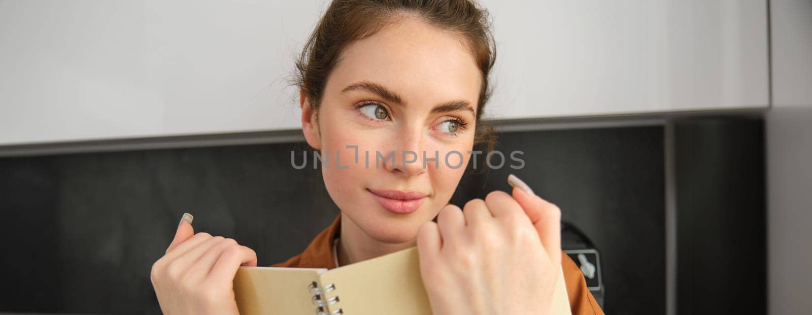 Close up portrait of young romantic woman, smiling, looking aside, holding notebook, reading notes, writing in diary, checking her planner.