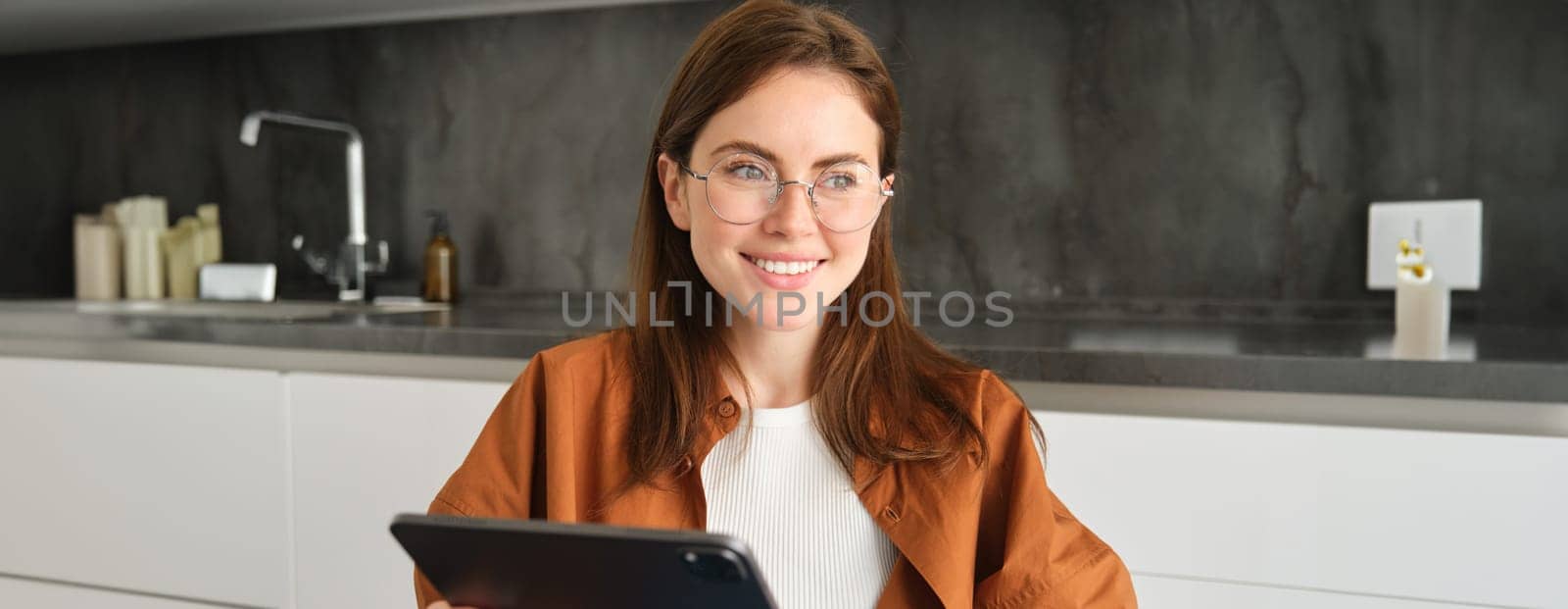 Portrait of young beautiful woman in glasses, working from home, studying online in her kitchen, holding digital tablet, online shopping.