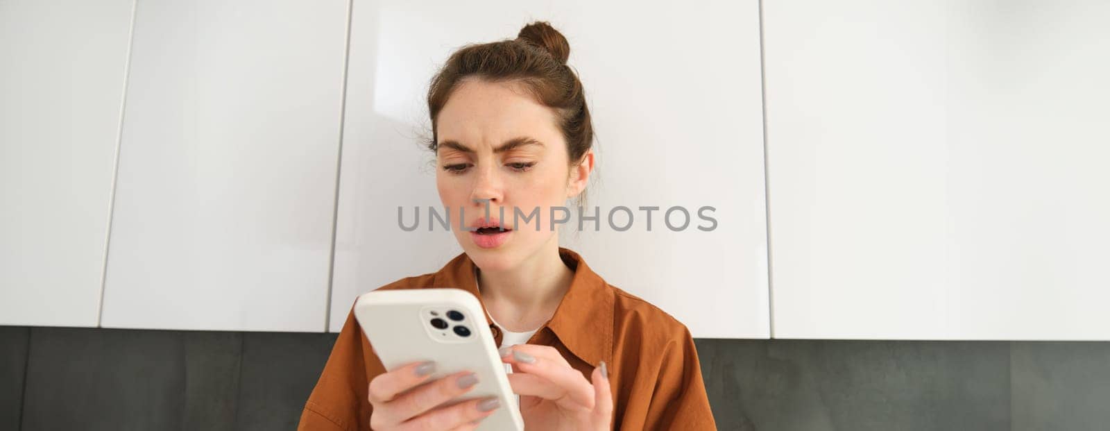 Portrait of woman with frowned face, looking at her mobile phone screen, scrolling social media feed, buying online, shopping on an app by Benzoix