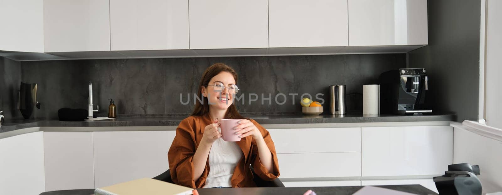 Portrait of young woman working from home, taking a break for cup of tea. Girl studies remotely, sits in kitchen, drinking hot coffee and smiles.
