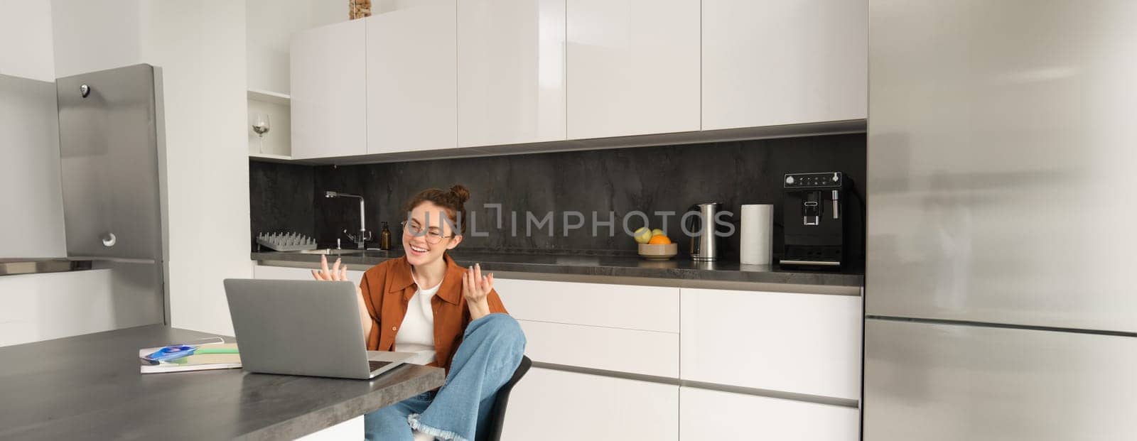 Portrait of young stylish woman working from home, sitting in kitchen with laptop, joining online meeting and talking to client on remote, video chats on computer.